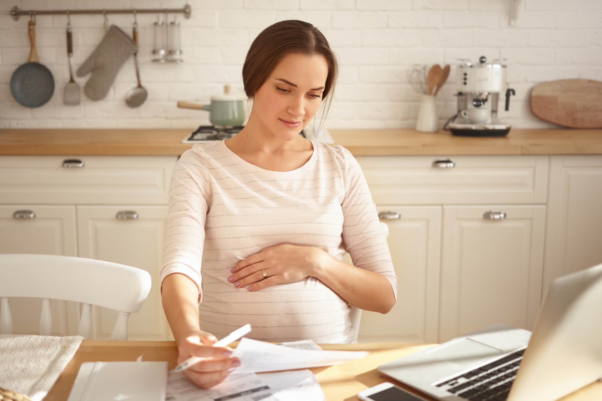 Picture of attractive young brunette expectant mother sitting in kitchen in front of open laptop, surrounded with bills, doing paperwork and calculating finances, holding pen and touching belly