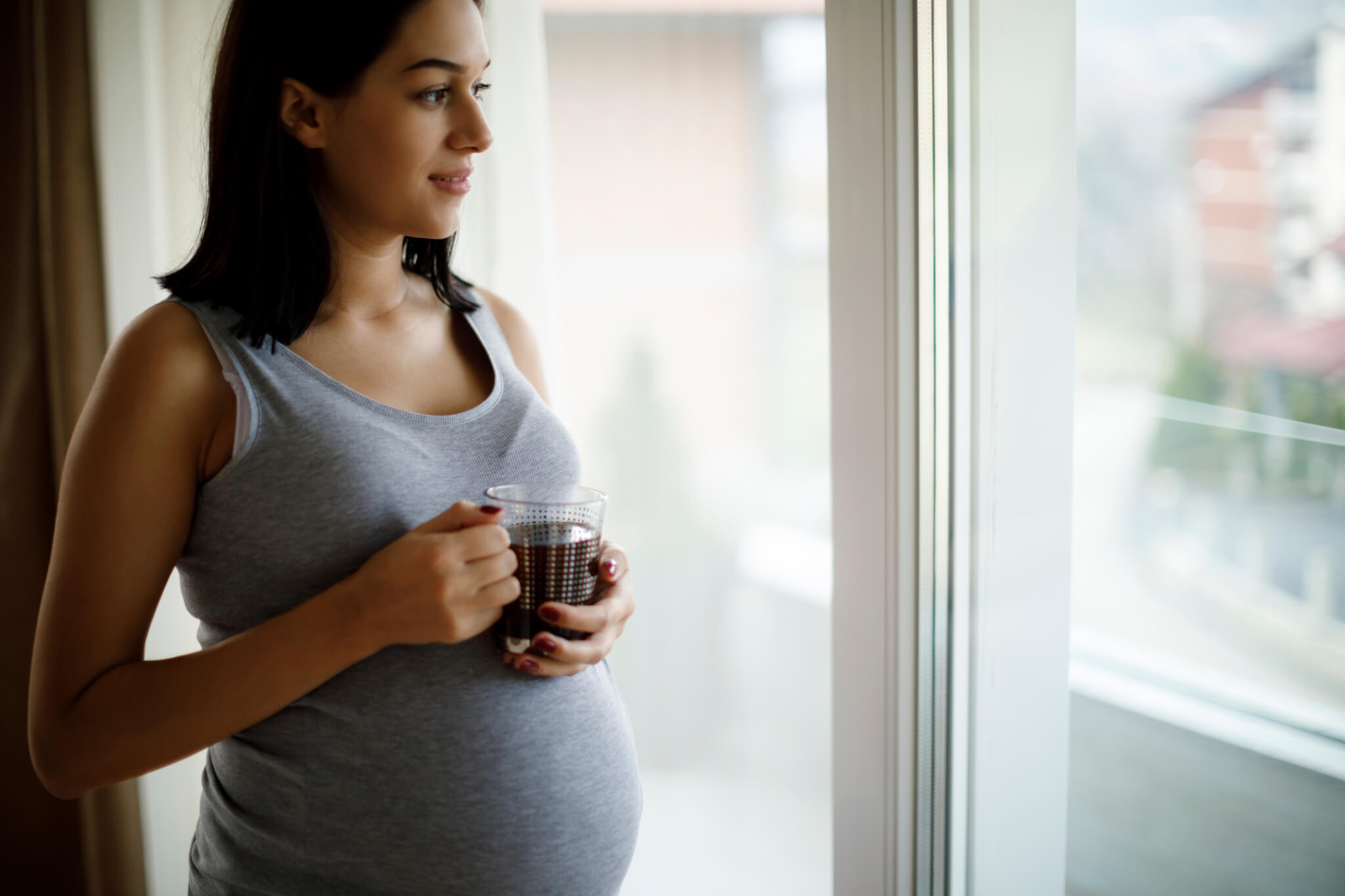 Portrait of young happy pregnant woman standing by the window and drinking tea